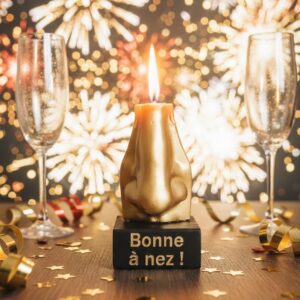 Bougie insolite nez doré allumée “Bonne à nez” sur socle noir avec flûtes de champagne et feux d’artifice en arrière-plan