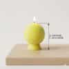 Une image présentant une bougie en cire, modelée en forme de balle de tennis, de couleur jaune fluo avec des lignes blanches. La bougie est allumée et posée sur un support en cire jaune fluo, sur une surface en bois clair, devant un fond uni blanc. Une flèche d'indication de hauteur est visible à droite, avec l'inscription "7 cm" et "HAUTEUR DE LA BASE".