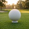 Photographie de produit d'une bougie blanche en forme de balle de golf, modélisée en 3D avec tous ses alvéoles, allumée et posée sur son socle. La bougie est au centre d'un green de golf en herbe, avec un filet de practice et des arbres flous en arrière-plan sous un éclairage doux de fin de journée. Une création artisanale unique de La Fée des Bougies.