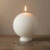 Photographie de produit d'une bougie blanche en forme de balle de golf, allumée, posée sur un socle blanc. Un indicateur de mesure en bas de l'image précise un diamètre de 12 cm. Fond neutre texturé et support en bois pour une ambiance élégante et artisanale.