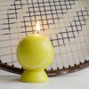 Photographie de produit d'une bougie en cire jaune lisse en forme de balle de tennis, avec une mèche allumée, posée devant une raquette de tennis en bois vintage. Ambiance chaleureuse, création artisanale La Fée des Bougies.
