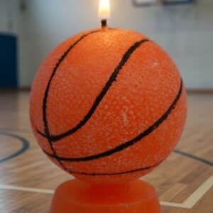 Une image présentant une bougie en cire, modelée en forme de ballon de basket, de couleur orange avec des lignes noires. La bougie est allumée et posée sur un support en cire orange, sur un fond de terrain de basket d'intérieur avec un panneau de basket visible en arrière-plan.
