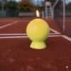 Une image présentant une bougie en cire, modelée en forme de ballon de tennis, de couleur jaune fluo avec des lignes blanches. La bougie est allumée et posée sur un support en cire jaune fluo, sur un fond de terrain de tennis d'extérieur en terre battue avec un filet, un banc et une chaise d'arbitre visibles en arrière-plan