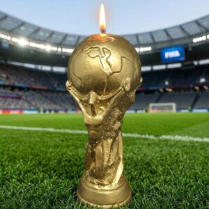 Photographie de produit d'une bougie dorée en forme de Trophée de football, allumée et posée sur la pelouse verte d'un stade mythique de l'équipe de France. Mèche enflammée, design réaliste des deux figures soutenant le globe, création artisanale exclusive de La Fée des Bougies.