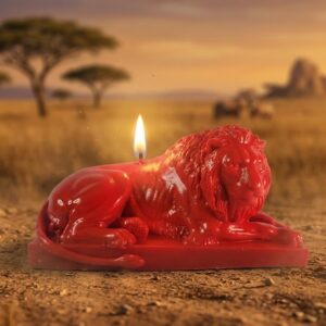 Photographie d'une bougie lion artisanale rouge et allumée, posée sur le sol sablonneux d'une savane africaine. Elle représente un lion mâle majestueux en repos. Moulage d'art La Fée des Bougies.