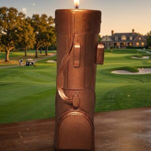 Photographie de produit d'une bougie en forme de sac de golf de couleur bronze, allumée avec une flamme vive. La bougie est posée sur un support en bois avec, en arrière-plan, un terrain de golf verdoyant et son club-house au coucher du soleil. Création artisanale La Fée des Bougies.