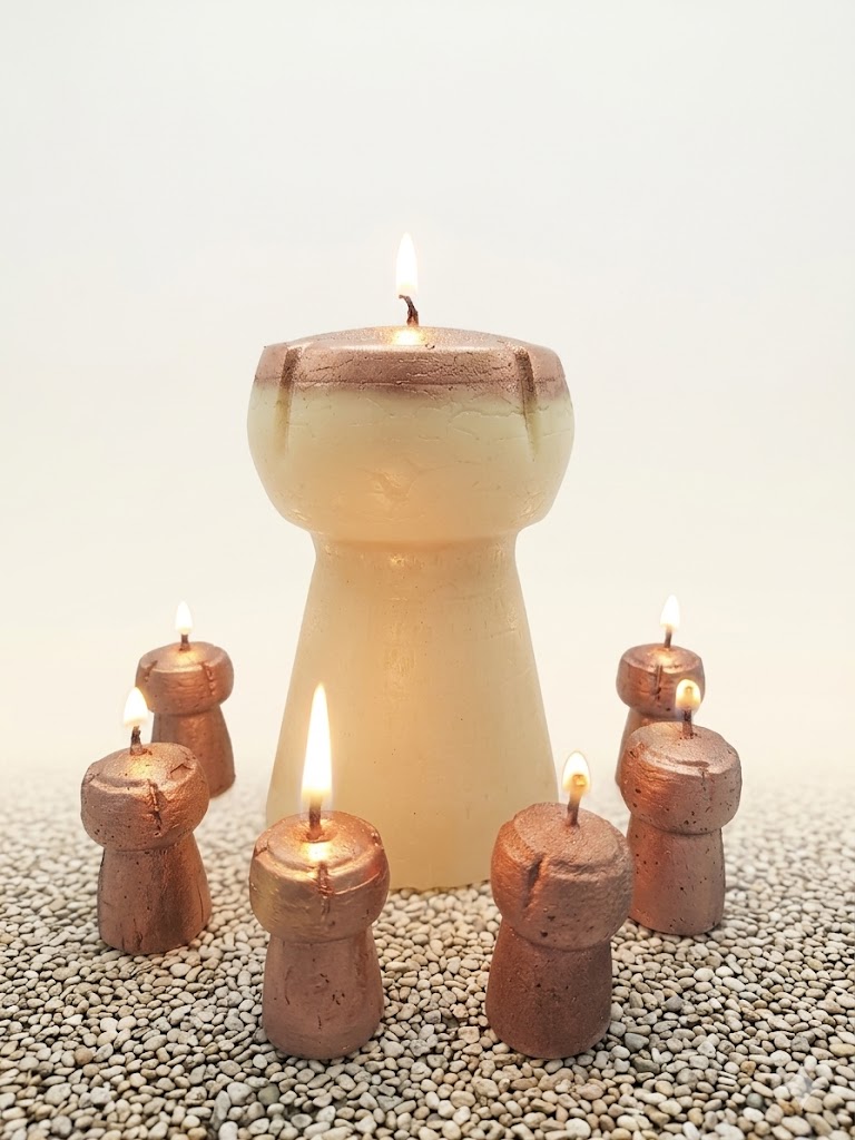 Ambiance chaleureuse avec le set exclusif de bougies bouchons de champagne allumées, comprenant le grand modèle de 14 cm et les six petits bouchons taille réelle, posés sur un lit de petits cailloux blancs.