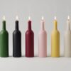 Alignement de six bougies artisanales en forme de bouteilles de Bordeaux (vert, noir, rouge, rose, jaune, blanc) sur fond neutre.